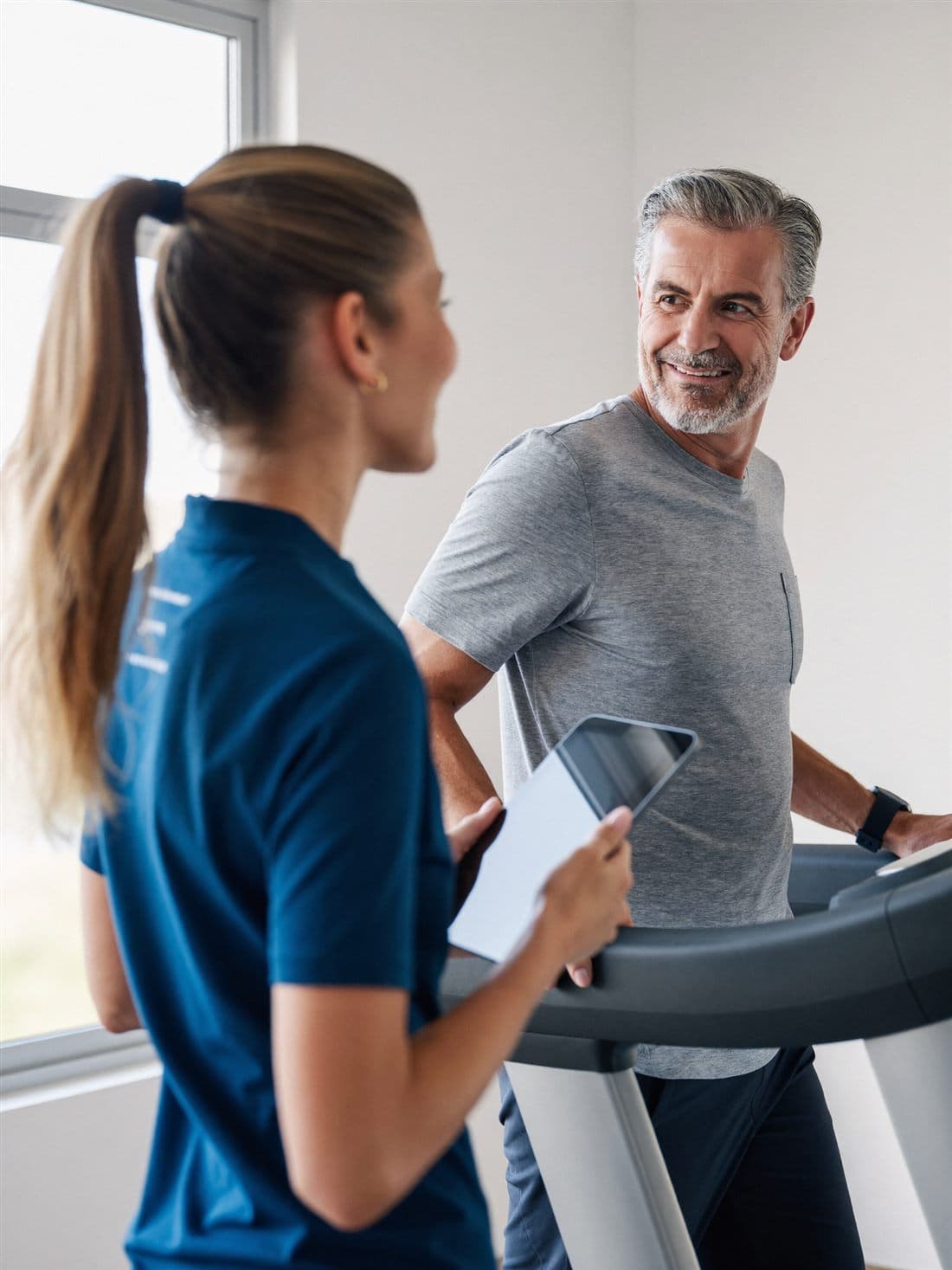 A cardiac exercise coach holds a tablet and encourages her client as he walks a monitored treadmill session in a bright private studio